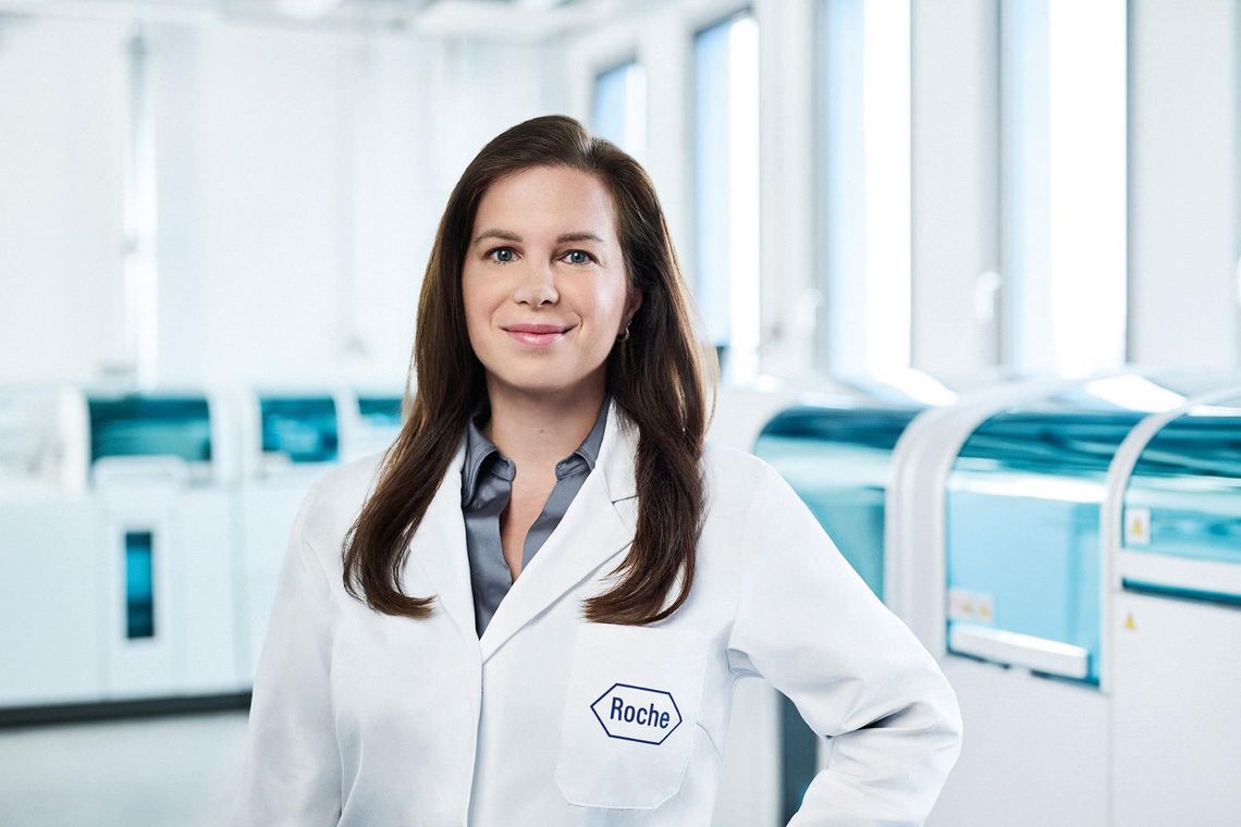 Frau im weißen Laborkittel mit Roche-Logo steht in einem modernen Diagnostiklabor mit Analysegeräten im Hintergrund.