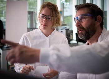Zwei Laborfachkräfte in Schutzkleidung und Schutzbrillen arbeiten gemeinsam im Labor und betrachten aufmerksam eine Versuchsanordnung.